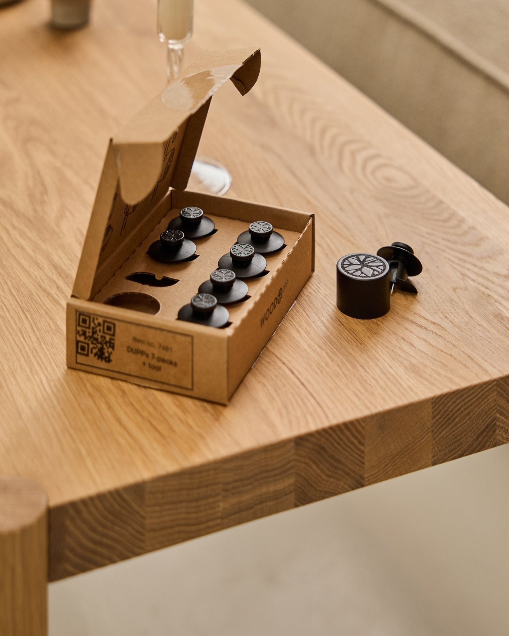 WoodUpp Dupps Original set (7 pieces and 1 tool) arranged on a wooden table, with one Dupps piece and part of a glass visible at the edge.